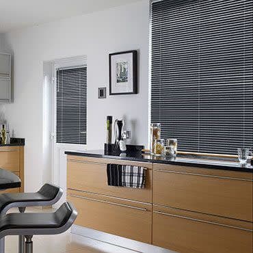 Grey horizontal Venetian blinds covering a large window and adjacent door window, slats tightly closed dimming light; in a modern kitchen with light-wood cabinets, black countertop, bar stools, small appliances.