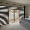 Zebra roller blinds with alternating dark and translucent horizontal stripes, partially lowered; curtains drawn right. Central sliding doors reveal a small balcony in a lit bedroom with grey bedding.