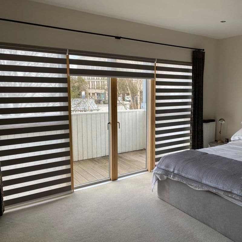 Zebra roller blinds with alternating dark and translucent horizontal stripes, partially lowered; curtains drawn right. Central sliding doors reveal a small balcony in a lit bedroom with grey bedding.