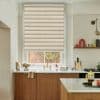 Layered beige zebra roller blind, lowered and filtering soft daylight across the window; modern kitchen context with wooden cabinets, white marble countertops, brass sink faucet and a brick-and-greenery view.