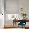 Light-grey Roman blind with a subtle circular pattern, lowered and softly filtering daylight above a desk with a teal chair, small mirror and vase of flowers in a neutral, sunlit study.