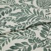 Botanical-printed cotton fabric, folded across the center revealing a layered edge, green leaf-and-fruit motifs on an off-white ground; close-up textile sample on a neutral surface under soft studio light.