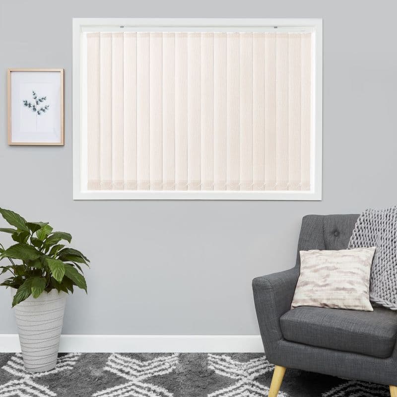 Beige vertical blinds fully closed over a white-framed window; diffusing soft daylight; in a neutral living room with grey walls, potted plant, framed print, grey armchair, patterned rug.