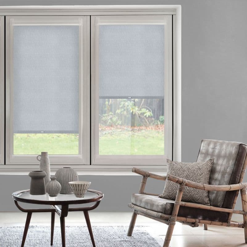 Two light-gray roller blinds covering adjacent windows, left fully lowered, right partially raised showing garden; light-filtering fabric with visible hem; modern living room with neutral tones and rattan chair.