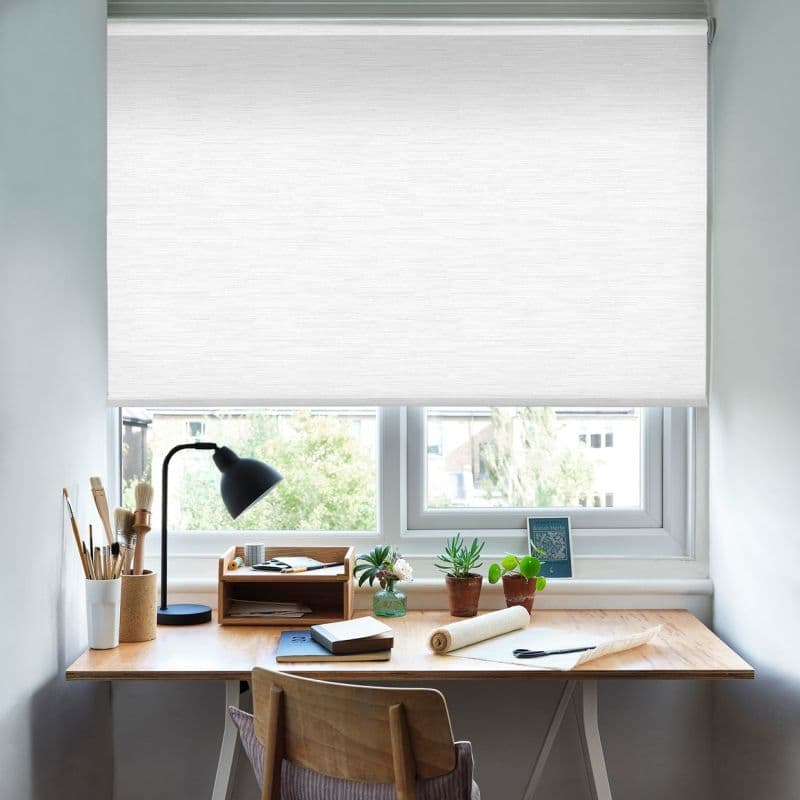 White textured roller blind lowered almost fully across the window, filtering soft daylight; wood desk beneath holds a lamp, brushes, notebooks and potted succulents in a calm home office/study.