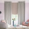 Floor-length pleated grey curtains frame a lowered beige Roman blind; drawn to the sides while the blind filters light, revealing a leafy view in a softly lit pastel living room.
THE FASHION BOOK