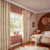 Cream grommet curtains with brown spots, hung on a brass rod, parted to frame and softly filter daylight through the window; warm pink bedroom with a brass bed and plants.