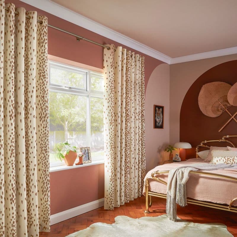 Cream grommet curtains with brown spots, hung on a brass rod, parted to frame and softly filter daylight through the window; warm pink bedroom with a brass bed and plants.