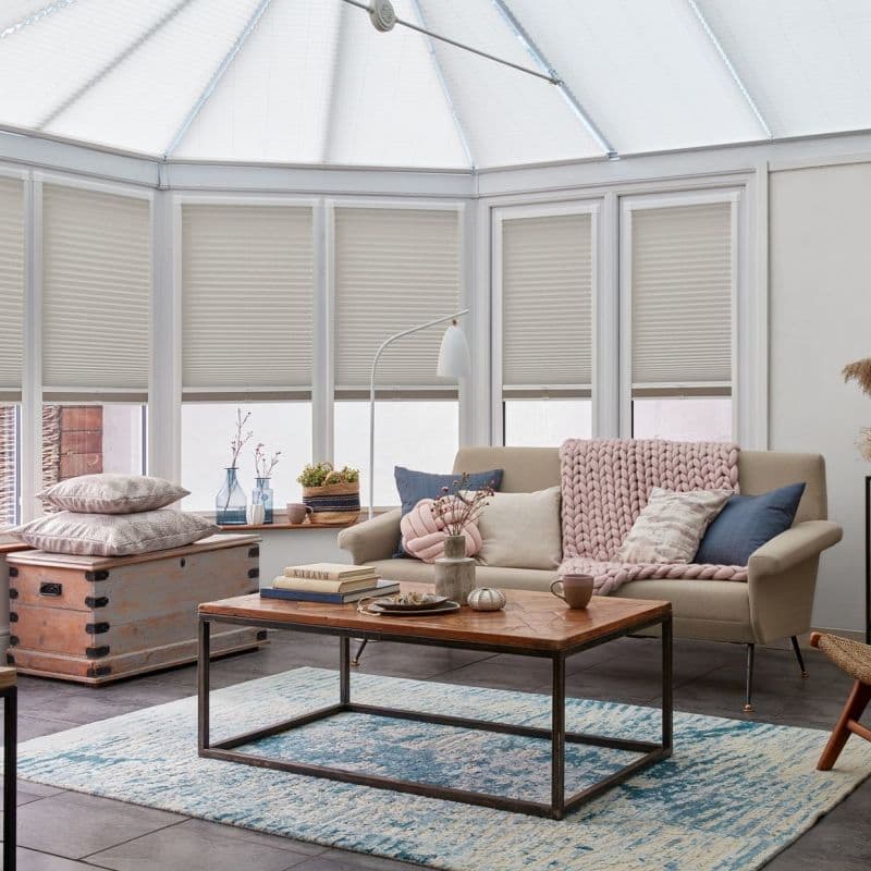Beige pleated cellular blinds, fitted inside tall bay/conservatory windows and partially lowered, diffusing daylight; cozy sunroom living area with neutral sofa, chunky pink throw, wooden coffee table and blue rug.