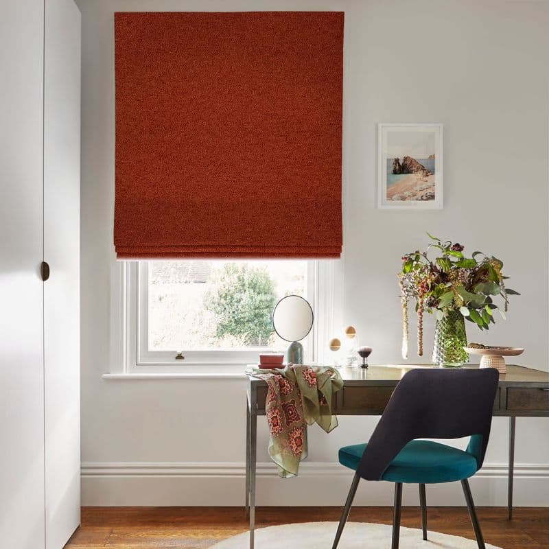 Rust-red roman blind lowered over a sash window, textured fabric with a folded bottom edge; blocking and softly diffusing daylight in a neutral, light-filled study with desk, teal chair, vase.