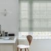 Sheer pale-gray Roman blind lowered over a multi-pane window, softly filtering daylight and revealing tree silhouettes; a minimalist home office with a white desk, wooden chair and pendant lamp.