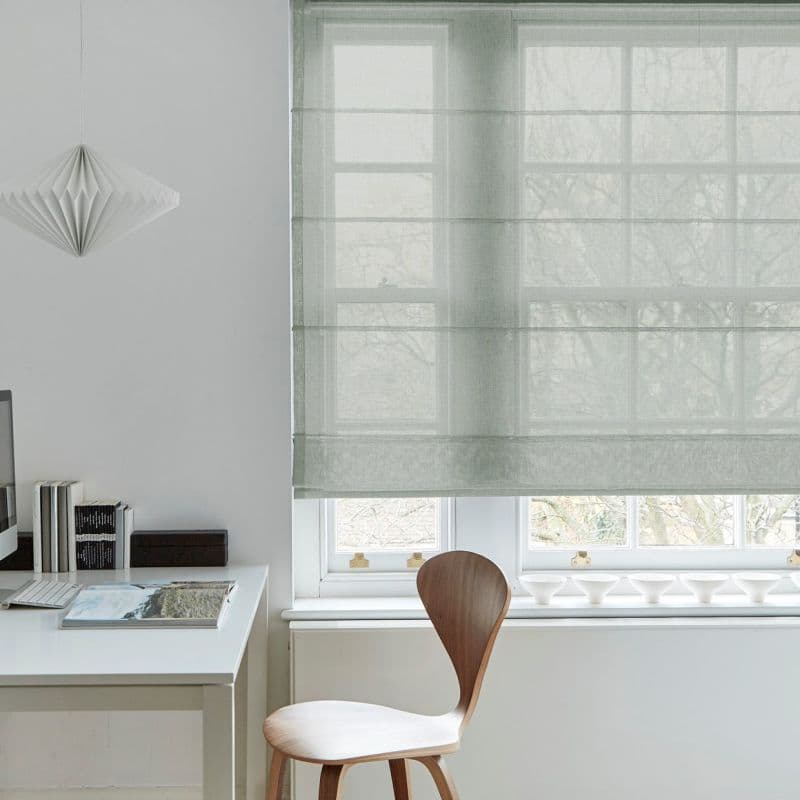Sheer pale-gray Roman blind lowered over a multi-pane window, softly filtering daylight and revealing tree silhouettes; a minimalist home office with a white desk, wooden chair and pendant lamp.