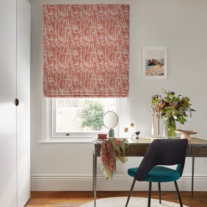 A terracotta, abstract-patterned Roman blind lowered over a sash window, softly filtering daylight; above a desk with a teal chair, vase of flowers, round mirror, and pale walls.