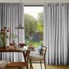Grey, floor-length pleated curtains framing a sliding glass door, slightly parted to admit soft daylight — wooden dining table set for breakfast with pancakes, pendant lamp, and garden view.