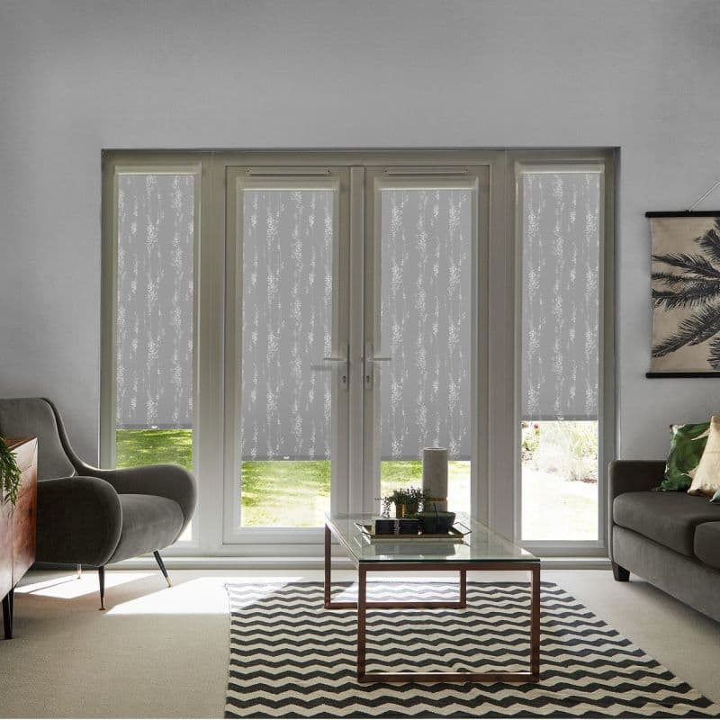 Patterned roller blinds (four panels) pulled down across French doors and sidelights, translucent vine motif filtering daylight; modern living room with grey sofas, glass coffee table and chevron rug.