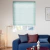 Light teal cellular blind, mostly lowered across a recessed window, softly filtering daylight; modern living room with a blue sofa, colored cushions, brass floor lamp and minimal art.