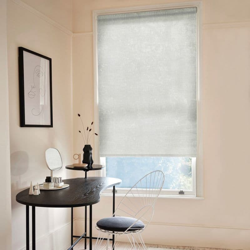 Light-gray patterned roller blind lowered, filtering soft daylight — over a single-pane window — in a pale, minimalist vanity corner with a black curvy desk, wire chair, and line-art wall print.
