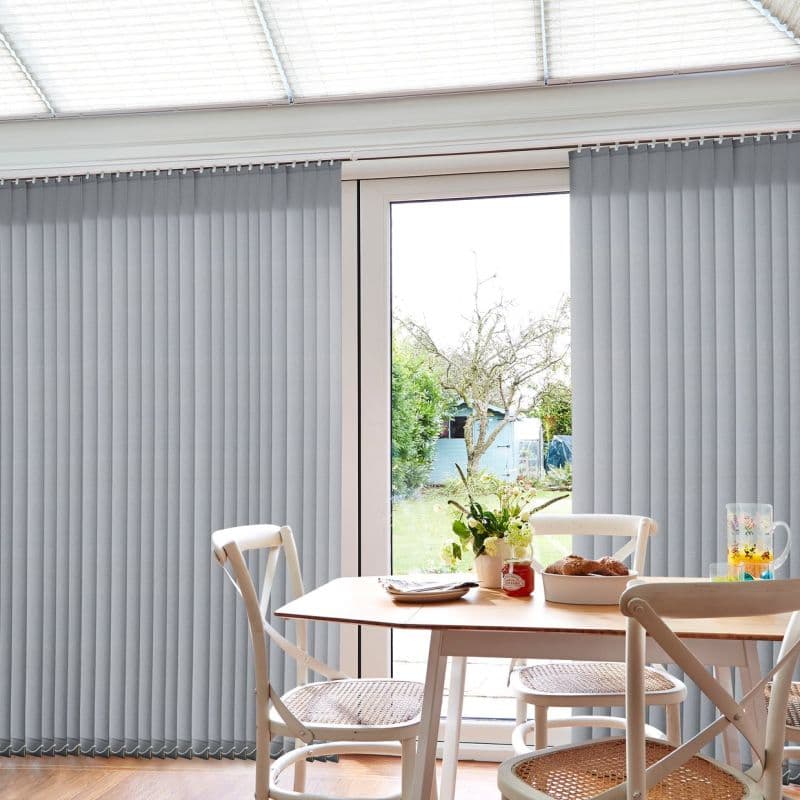 Grey vertical blinds span a sliding glass door, partially parted at center to reveal a garden; filtering daylight into a bright dining nook with wooden table, chairs, and plants.