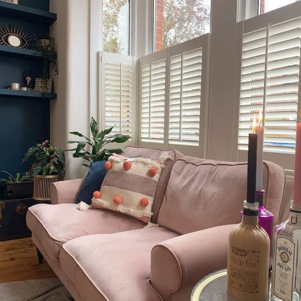White half-height louvered plantation shutters on bay windows, closed and filtering soft daylight; cozy living room with a pink velvet sofa, pom‑pom cushion, houseplants, dark shelving and candles.