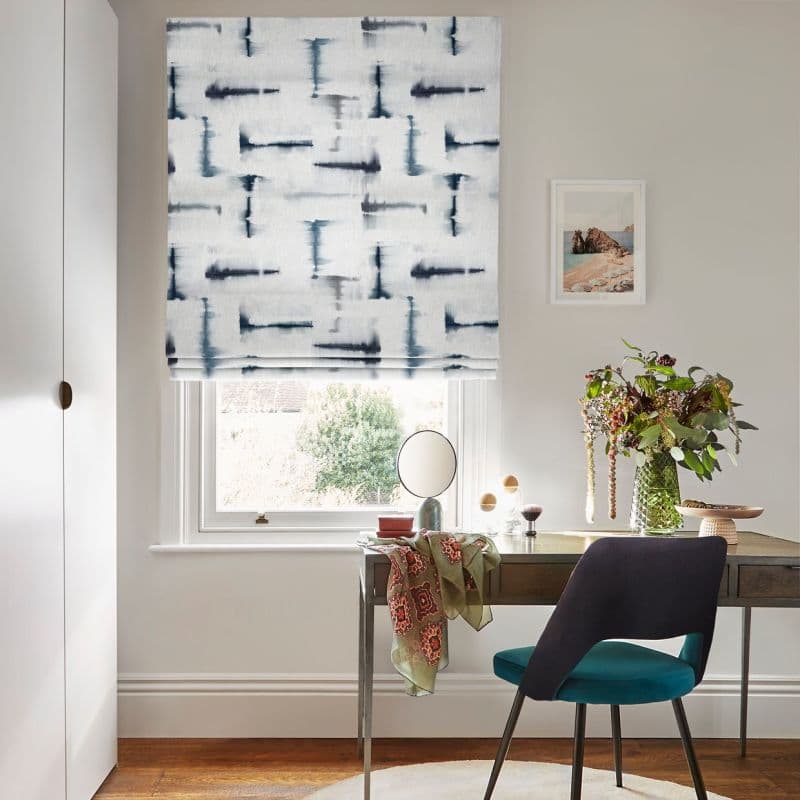 Roman blind with white fabric and navy abstract brushstrokes, lowered and folded at window, filtering soft daylight over a desk and teal chair in a tidy home office with vase of flowers.