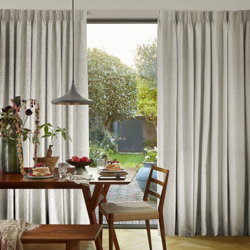 Floor-length pleated beige curtains, neatly pleated and drawn aside at the center, revealing a sunlit garden; dining table with wooden chairs, pendant lamp, and breakfast plates in the foreground.