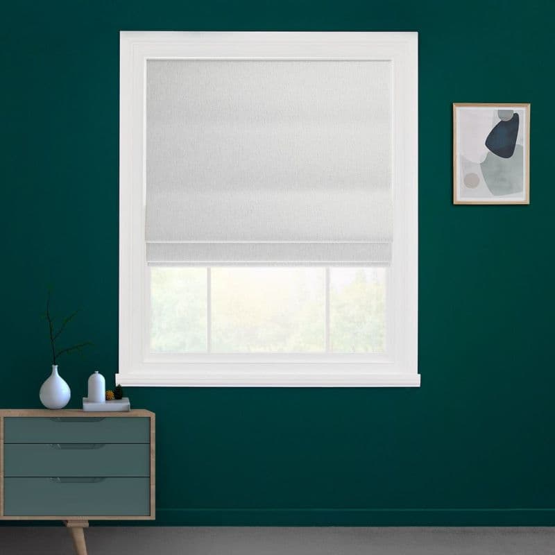 White fabric Roman shade, lowered to mid-window with horizontal folds, filtering soft daylight through lower glass — set in a teal-walled room with white trim, small dresser and framed abstract art.