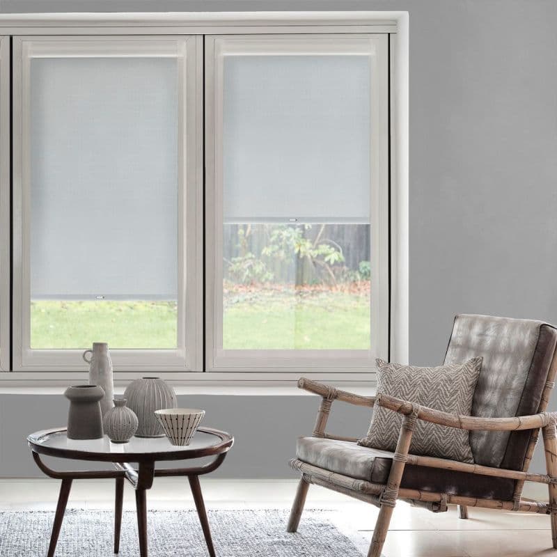 Two white roller blinds lowered mid-height, semi-translucent, filtering daylight; gray-walled living room with rattan leather armchair, patterned cushion, small round coffee table with ceramic vases, grassy yard beyond.