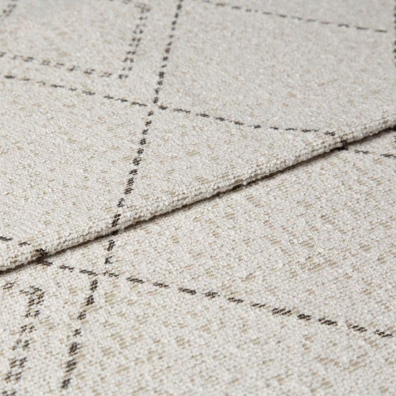 Beige woven rug with thin dashed dark grid lines, folded at the edge to show layered thickness and looped texture; close-up studio shot with neutral lighting on a flat surface.