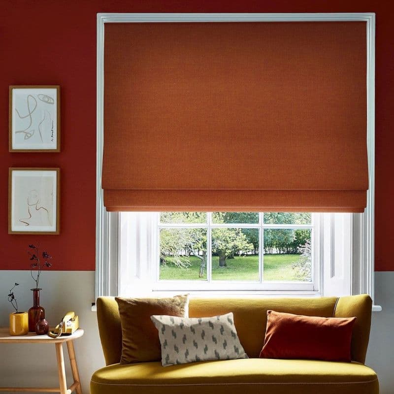 Burnt-orange Roman blind, lowered to the sill with soft folds; blocking light and framing a white-paned window; living room with mustard sofa, cushions, red walls, framed sketches and garden view.