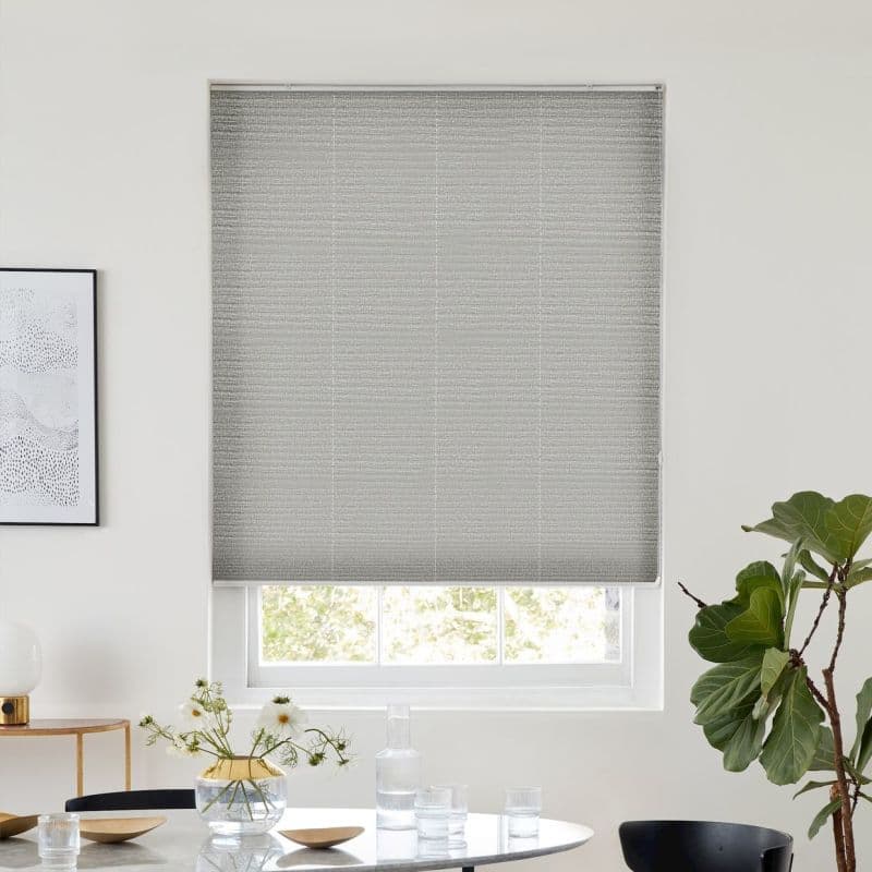 Grey textured roller blind fully lowered, blocking most light; minimalist dining nook with round table, glassware and vase, potted fiddle‑leaf fig, soft daylight filtering around blind edges.