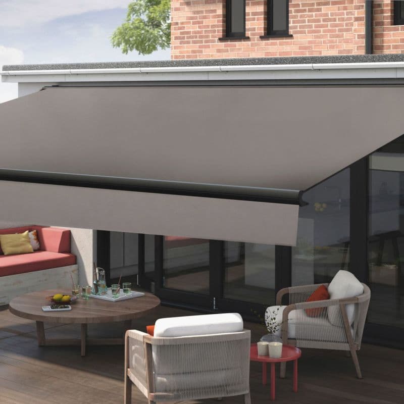 Retractable taupe fabric awning with black bar, fully extended over patio; shading and diffusing daylight across wooden deck. Modern home exterior with glass doors, brick upper wall, outdoor seating.