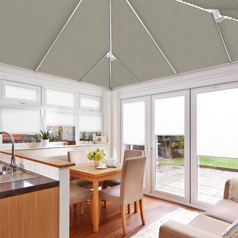 Grey pleated roof blinds with white cords, fitted across the conservatory ceiling; closed, filtering daylight over a bright kitchen-dining area with white-framed doors and windows opening to a patio garden.