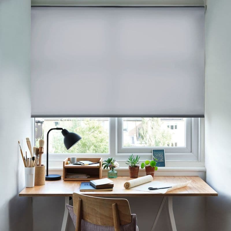 A white roller blind, lowered to cover most of the window and diffuse incoming light; a neat wooden desk beneath holds a lamp, potted plants, art brushes, notebooks, and a suburban daylight view.