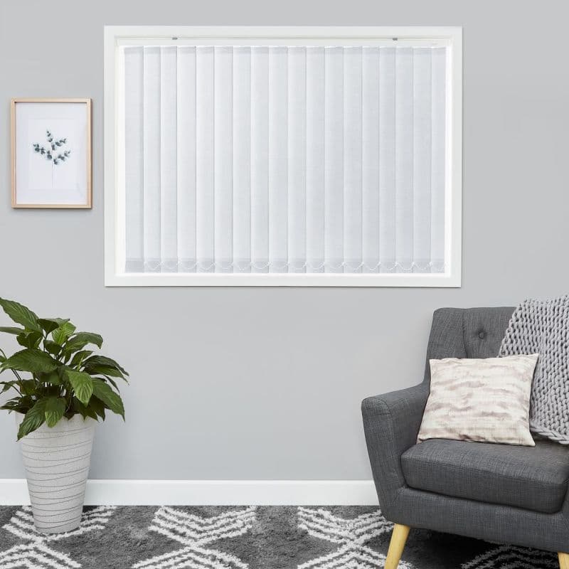 White vertical blinds covering a window, narrow fabric slats with scalloped bottoms; closed and diffusing soft daylight; living-room setting with grey armchair, knitted throw, potted plant, framed art.