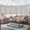 Beige pleated cellular blinds across tall conservatory windows, lowered and filtering soft daylight; a cozy sitting area with beige sofa, chunky pink throw, wooden coffee table and blue-pattern rug.