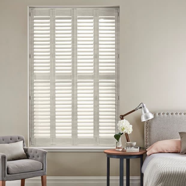 White plantation shutters—four tall louvered panels—fully closed, softly filtering daylight into a neutral bedroom corner with upholstered headboard, bedside table, lamp, vase of flowers and a gray armchair.