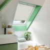 White pleated skylight blind, partially lowered over a slanted roof window, filtering daylight; set within a mint-green framed attic bathroom with white herringbone tiles, sink, tub, plant, and toiletries.