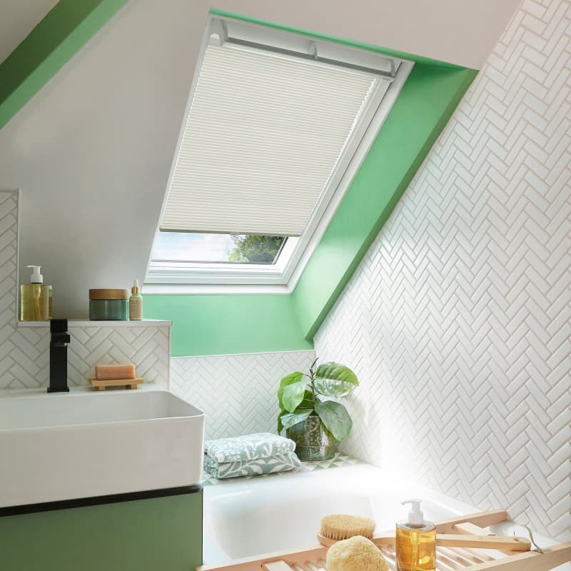 White pleated skylight blind, partially lowered over a slanted roof window, filtering daylight; set within a mint-green framed attic bathroom with white herringbone tiles, sink, tub, plant, and toiletries.