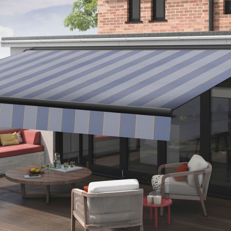 Striped retractable fabric awning with blue-gray diagonal stripes, fully extended over a patio; shading glass doors and outdoor seating — daytime, brick upper facade, wooden deck, lounge chairs and coffee table.
