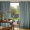 Teal-and-white patterned pleated curtains, hung on either side and slightly drawn to frame a central door, filtering daylight into a dining room with wooden table, chairs and garden view.