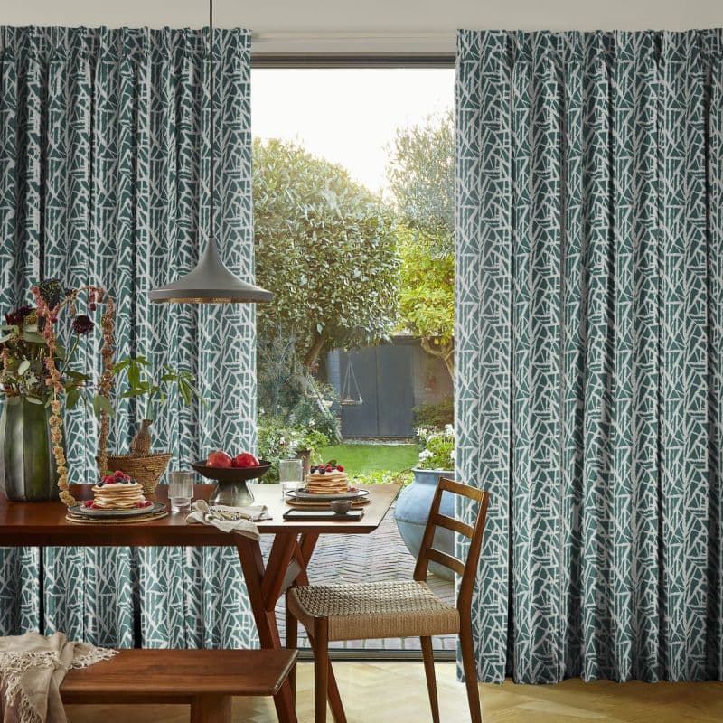 Teal-and-white patterned pleated curtains, hung on either side and slightly drawn to frame a central door, filtering daylight into a dining room with wooden table, chairs and garden view.