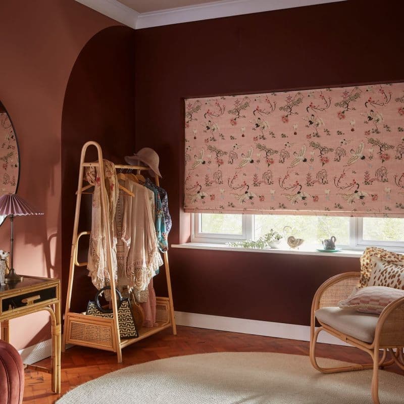 Pink fabric Roman blind with bird-and-floral print, fully lowered and gently diffusing daylight onto a cosy dressing corner with rattan clothing rack, soft seating, warm terracotta walls and parquet floor.