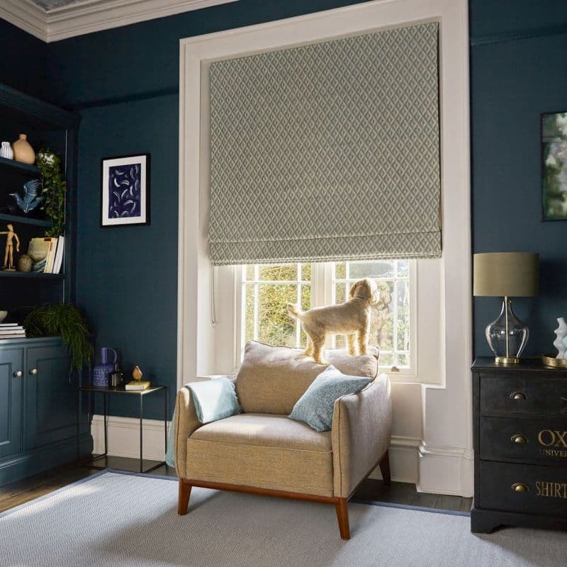 Patterned Roman blind with diamond motif, drawn almost fully down leaving a small gap; it filters soft daylight. Context: cozy teal living room, armchair by window with a small dog on the cushion.
OXFORD UNIVERSITY SHIRTS