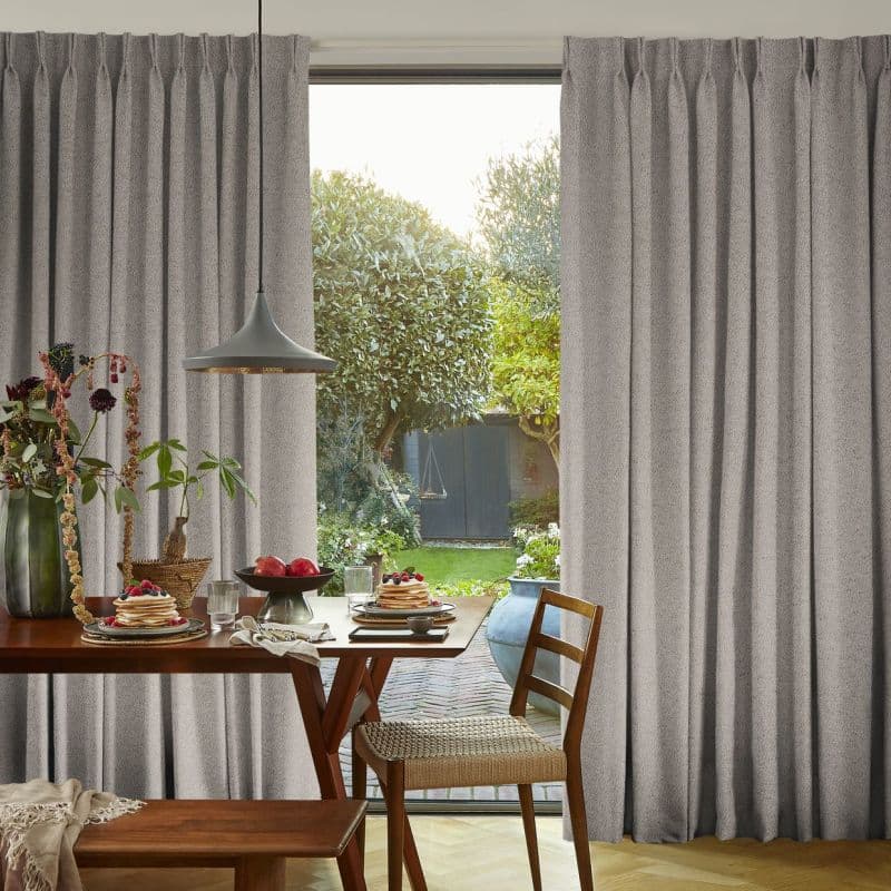 Grey pleated floor-length curtains, pulled aside to reveal a sliding glass door, framing a sunlit backyard; a wooden dining table with pancakes and a hanging pendant lamp fills the interior.