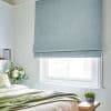 A pale blue Roman shade, lowered with horizontal folds, covers the window, filtering daylight; bedroom with white walls, green bedding, vase of white flowers and a small succulent.