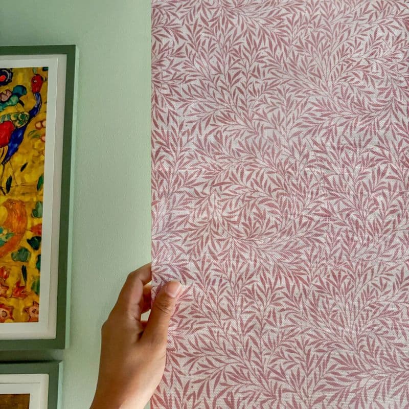 Pink fabric blind with white leaf pattern, held at its left edge by a hand; pale green interior wall with framed colorful artwork, softly lit.