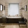 White horizontal venetian blinds fully lowered over center window, slats closed and filtering soft daylight; set above a built-in wooden bathtub in a neutral-striped, softly lit bathroom with sink, shelf, chair.