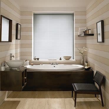White horizontal venetian blinds fully lowered over center window, slats closed and filtering soft daylight; set above a built-in wooden bathtub in a neutral-striped, softly lit bathroom with sink, shelf, chair.