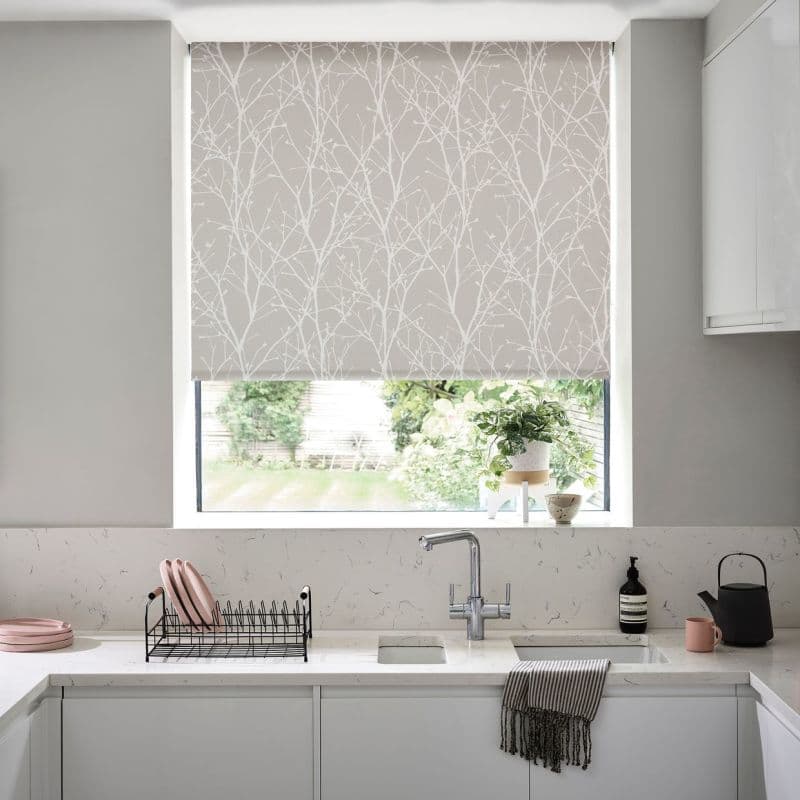 Patterned roller blind with white twig design, lowered to cover most of the window and filter daylight; modern kitchen sink with marble countertop, dish rack, potted plant and garden view.