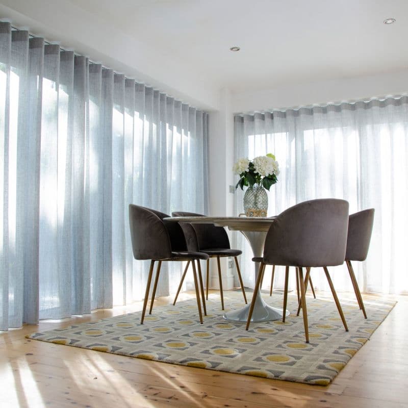 Object: sheer floor-to-ceiling pleated curtains on two walls, pale gray with vertical folds. Action: drawn closed, softly filtering daylight. Context: sunlit dining nook with round table, four chairs, patterned rug.